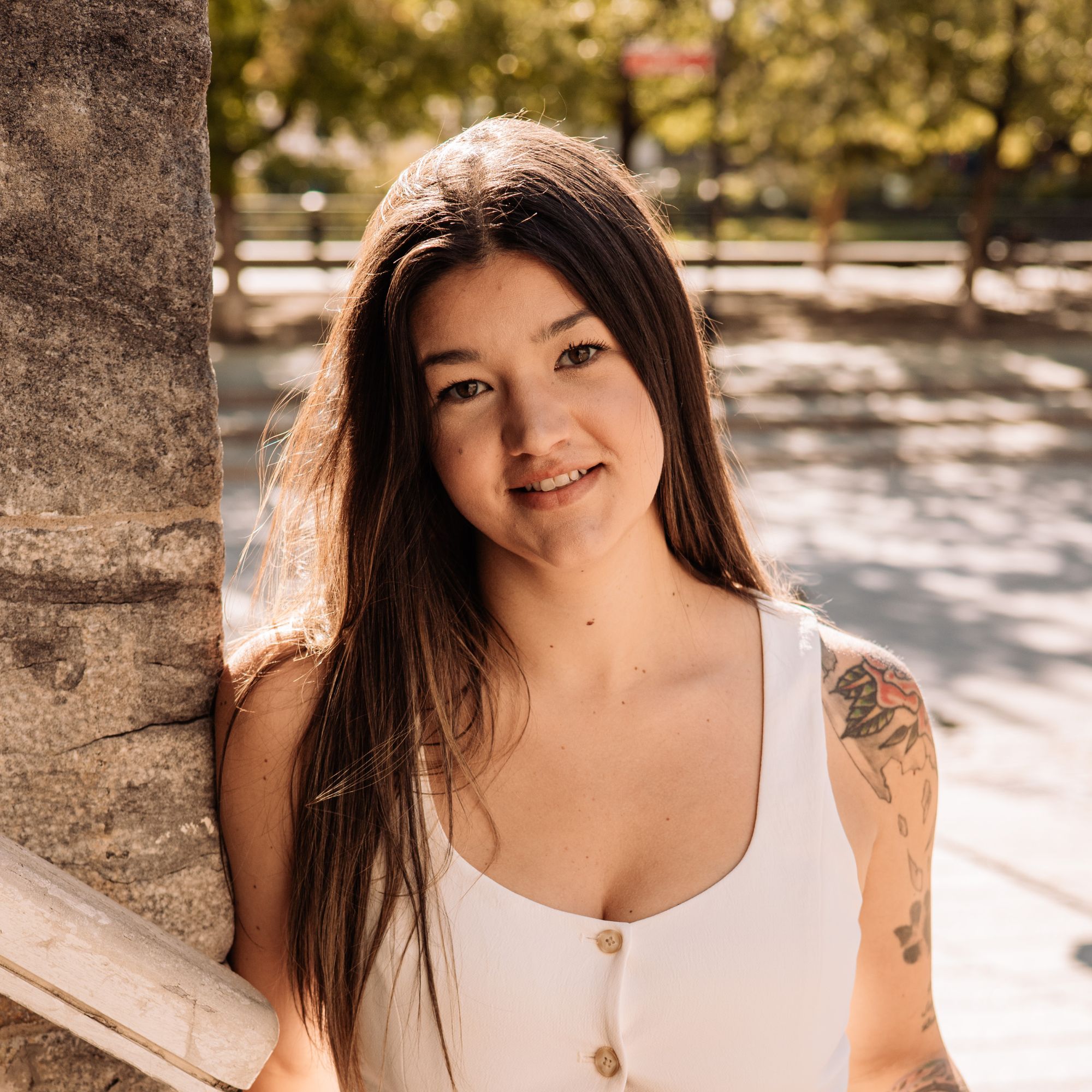 Smiling woman leaning against a textured tree trunk outdoors on a sunny day, partial face visible.