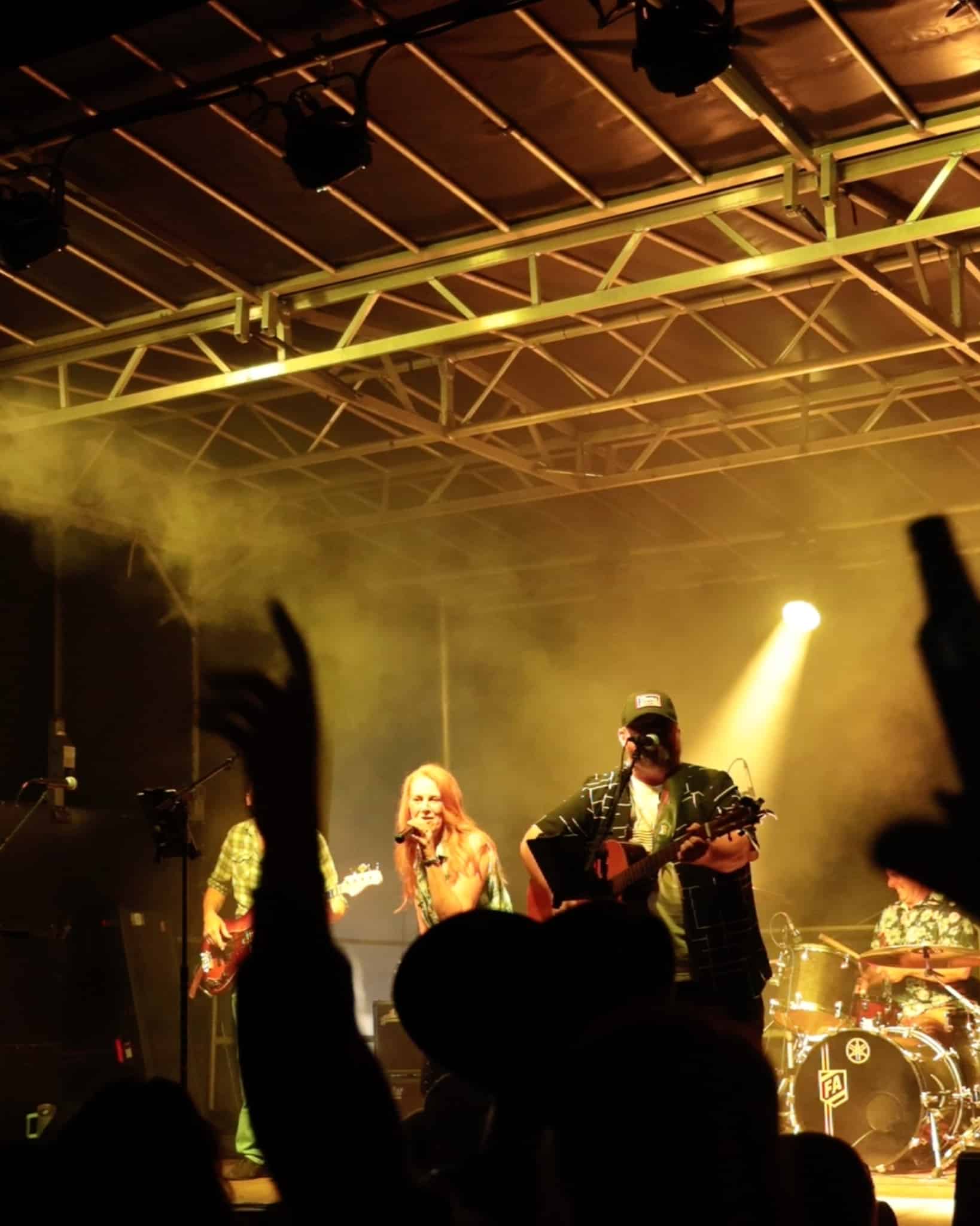 Band performs on a smoky stage with a female singer at center and guitarists on either side, audience silhouettes in front.
