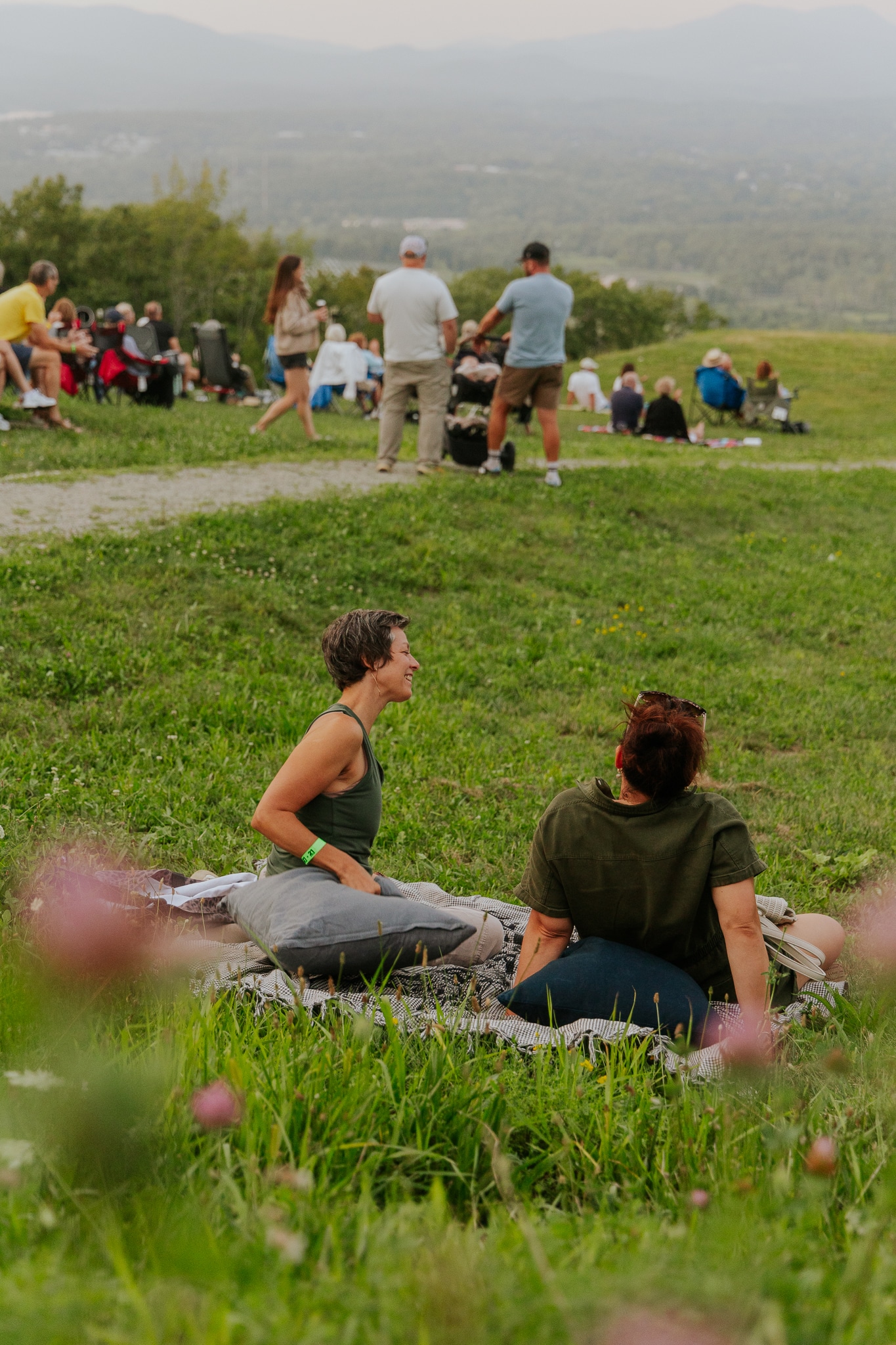 Two women chatting on a blanket in a grassy hilltop while a group picnics in the background.
