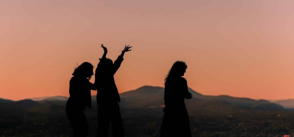 Three people stand in silhouette on a hill at sunset, arms raised against an orange sky with distant mountains in view.