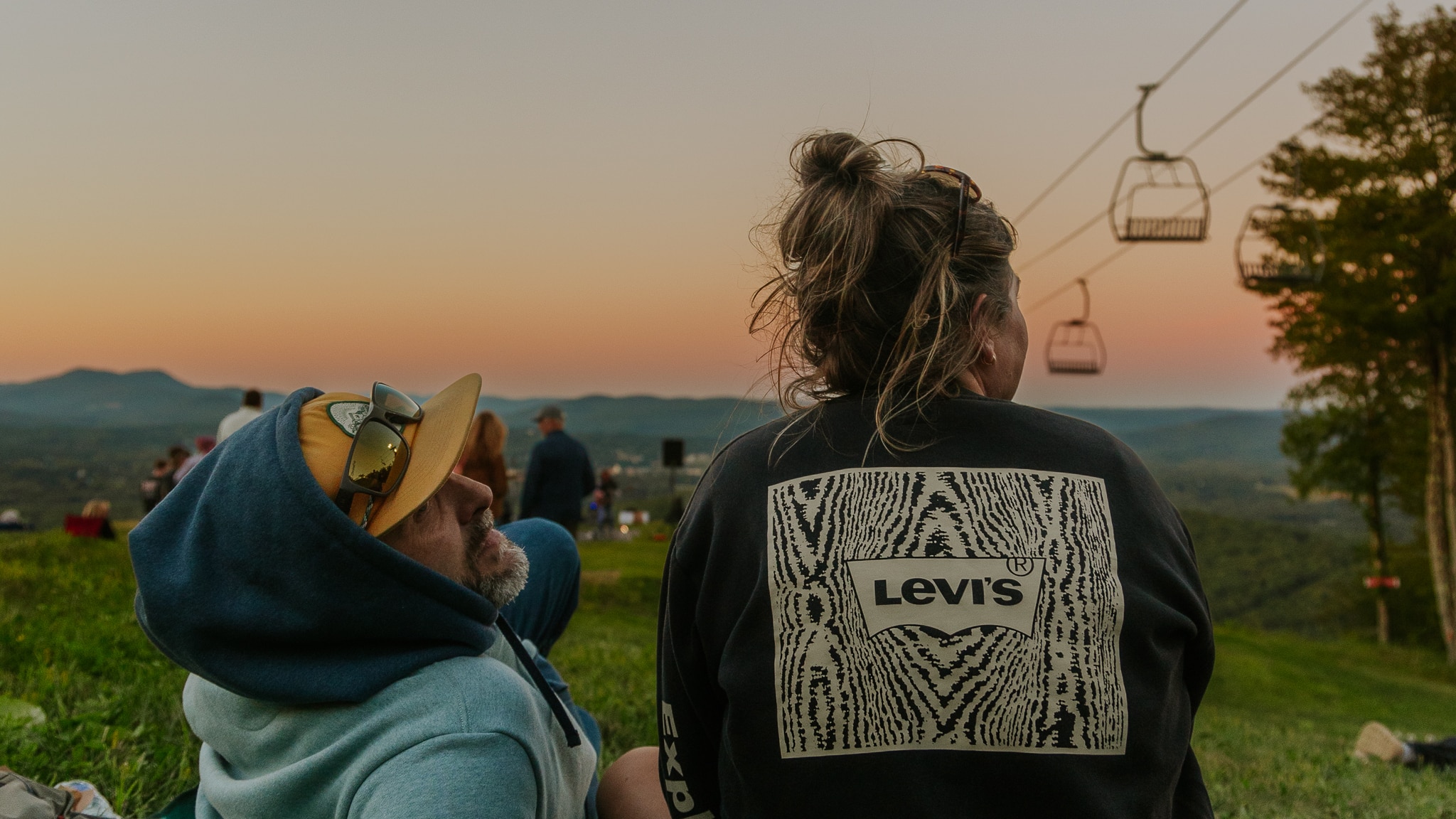 People relaxing on a grassy hill at sunset with chairlifts in the background; one person wears a Levi's sweatshirt visible from behind.