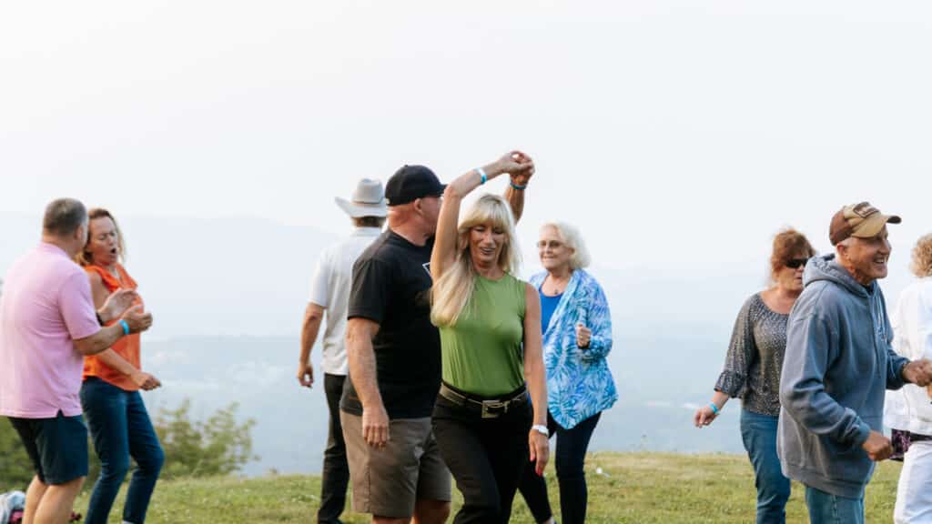 Group of adults dancing hand-in-hand on a hilltop with a scenic valley in the background, smiling.
