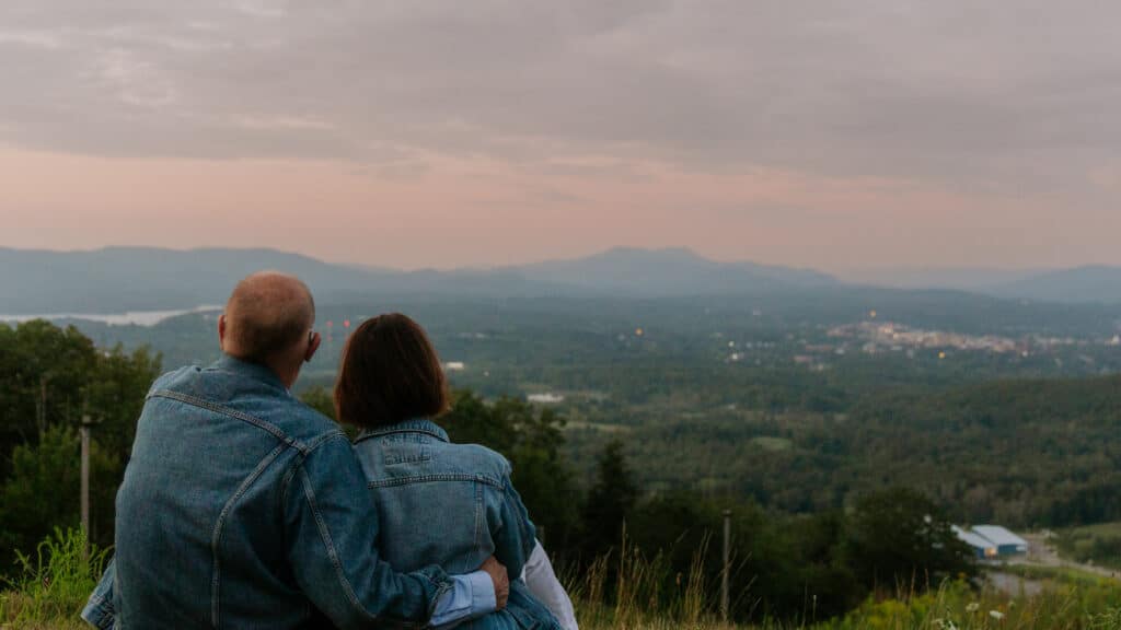 Couple in denim jackets sitting on a grassy hillside, cuddled together and looking out over a vast valley with distant mountains at sunset.