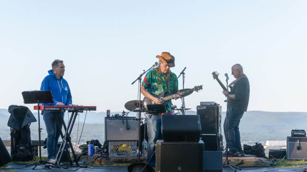 Three musicians perform on an outdoor stage with the keyboardist on the left, guitarist in the center, and bassist on the right, mountains in the background.