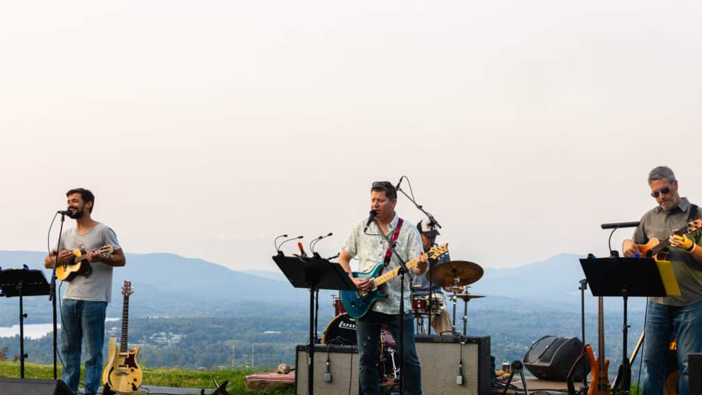 Band performing outdoors with three guitarists and drums against mountain backdrop, sunset sky behind them, microphones and stands visible, crowd off-frame.