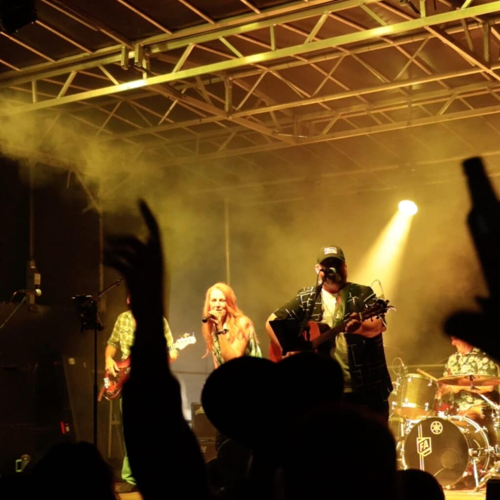 Live band performs on stage with guitarists and a vocalist, yellow stage lights and smoke, crowd silhouettes with raised hands in the foreground