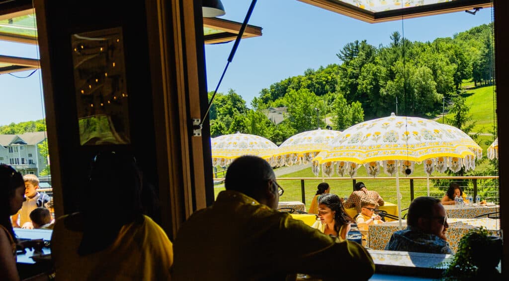 Outdoor cafe scene with people seated at tables under yellow floral parasols on a sunny day, hillside and trees in the background.