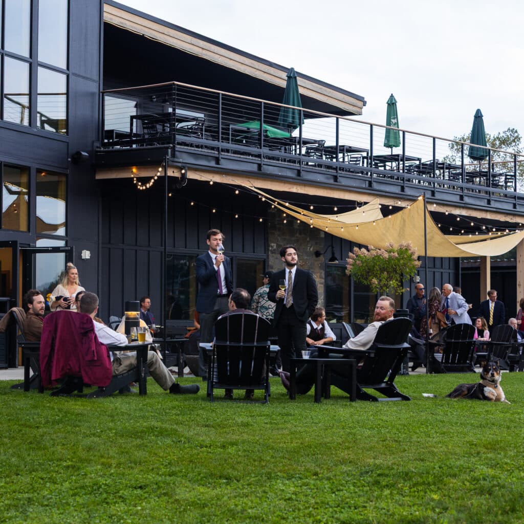 Guests in formal attire mingle on a patio with string lights and umbrellas, overlooking a dark building façade and green lawn with a dog at the right edge.