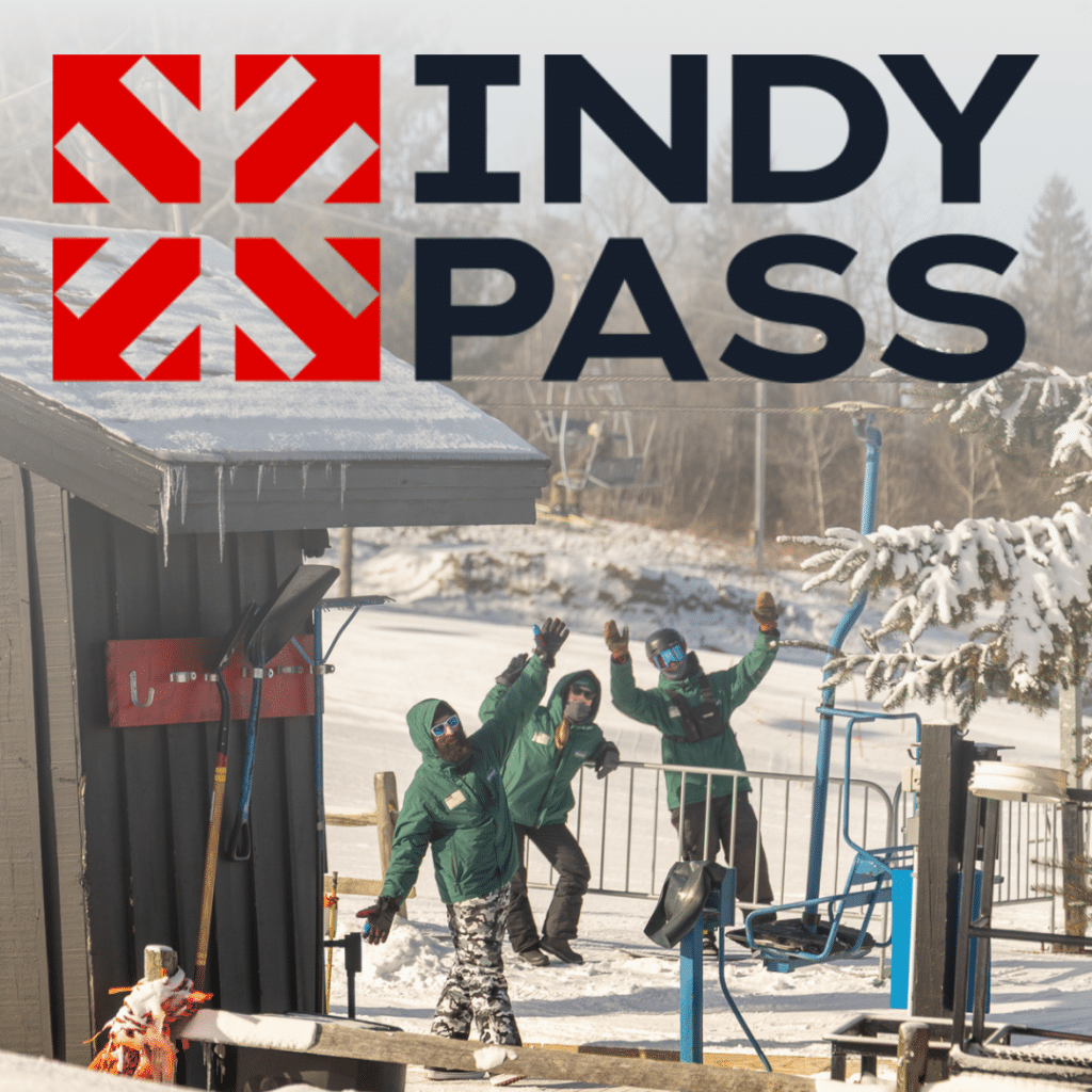 Indy Pass logo in bold black letters with a red geometric emblem, over a snowy ski area where four staff in green jackets wave at the camera.