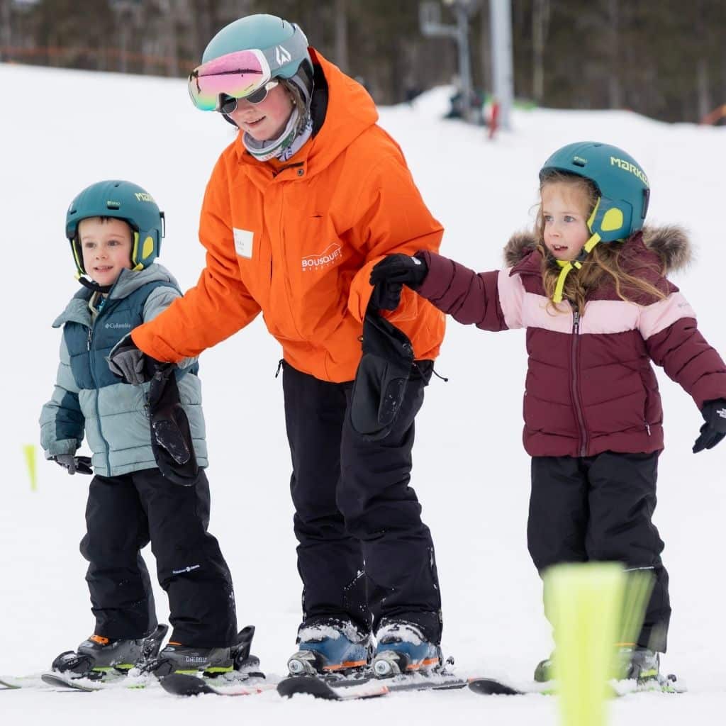 Instructor in an orange jacket guiding two children on skis, each wearing helmet and winter gear, holding hands as they practice on the snowy slope.