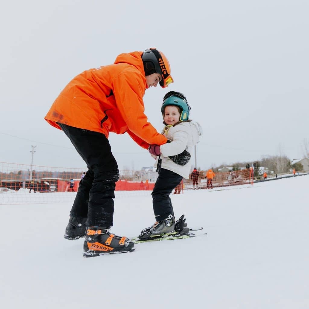Adult in an orange jacket helping a child on skis on a snowy slope, both wearing helmets and gear.