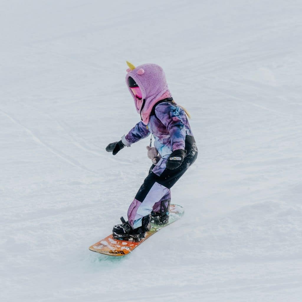 Child in a purple unicorn hooded jacket snowboards down a snowy slope wearing colorful gear and gloves.