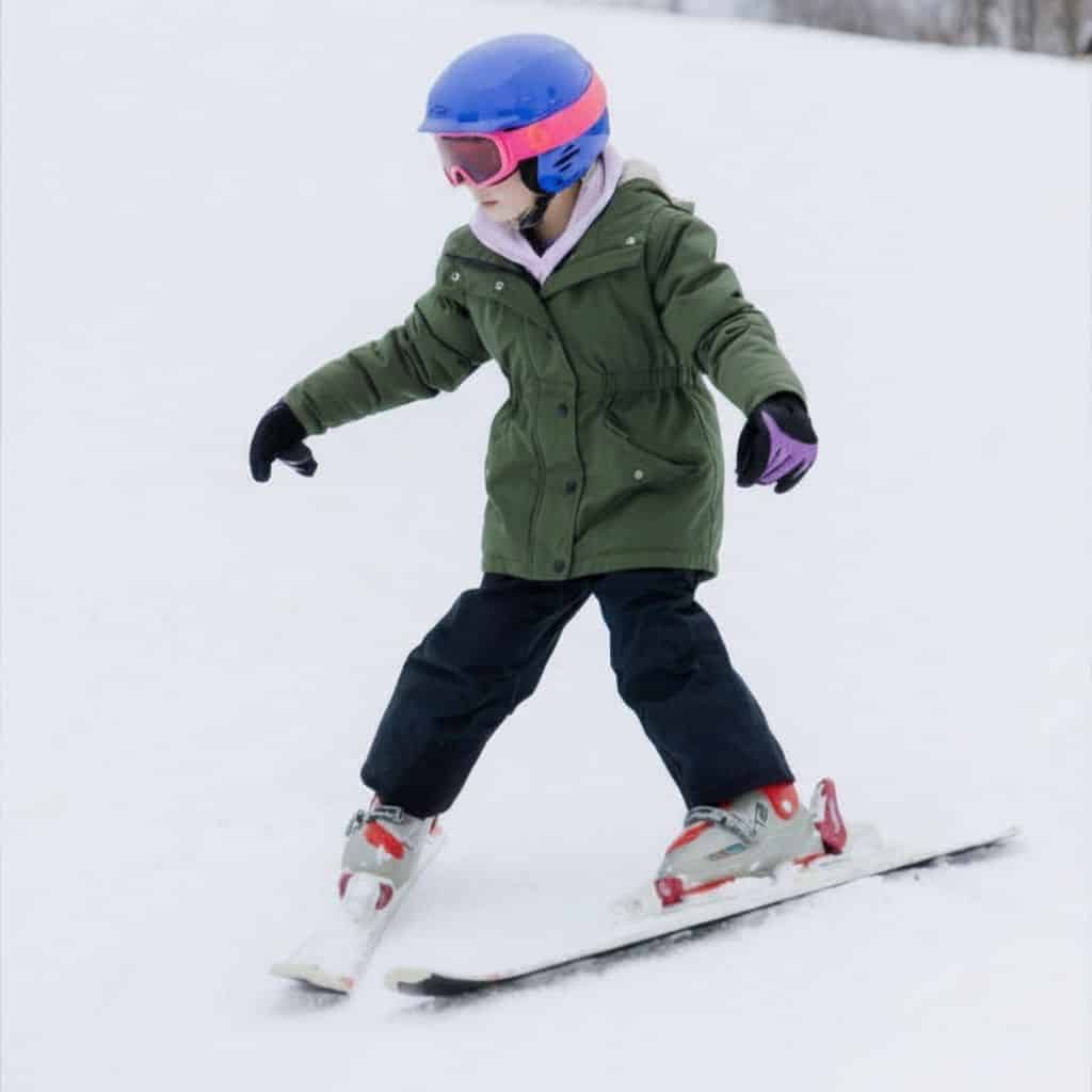 Child wearing a blue helmet and pink goggles skis downhill on a snowy slope in a green jacket and black pants with white skis and red bindings.