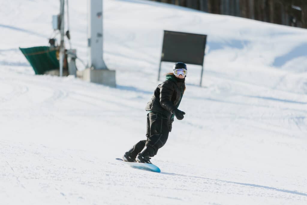 Snowboarder in a black jacket with a helmet and goggles carving down a sunny snowy slope, blue board visible.