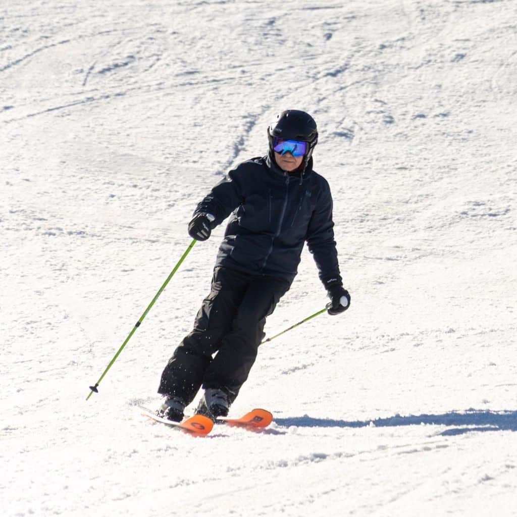 Adult skier in black gear wearing a helmet and reflective goggles carving down a snowy slope on orange skis with neon green poles.