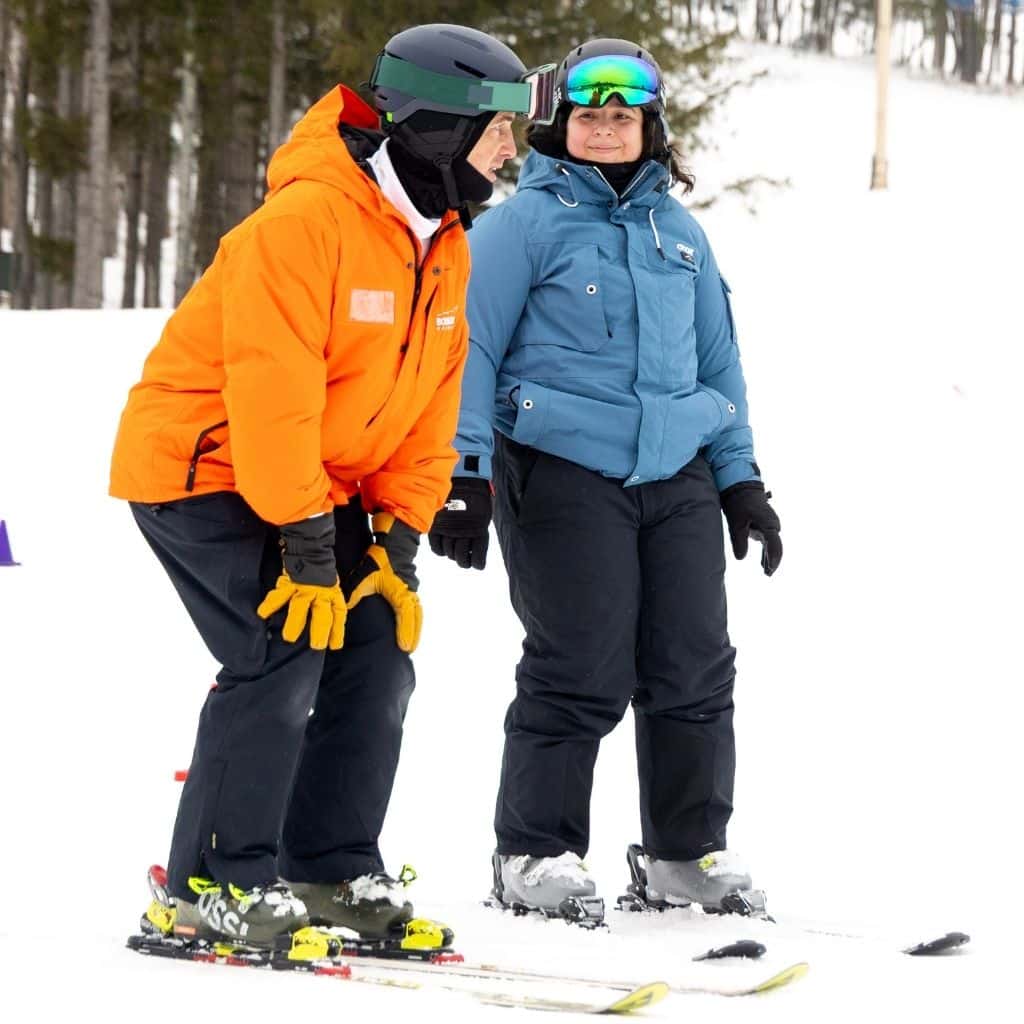 Two skiers in bright jackets stand on a snowy slope, chatting; one in orange with a helmet, the other in blue with goggles and gloves.