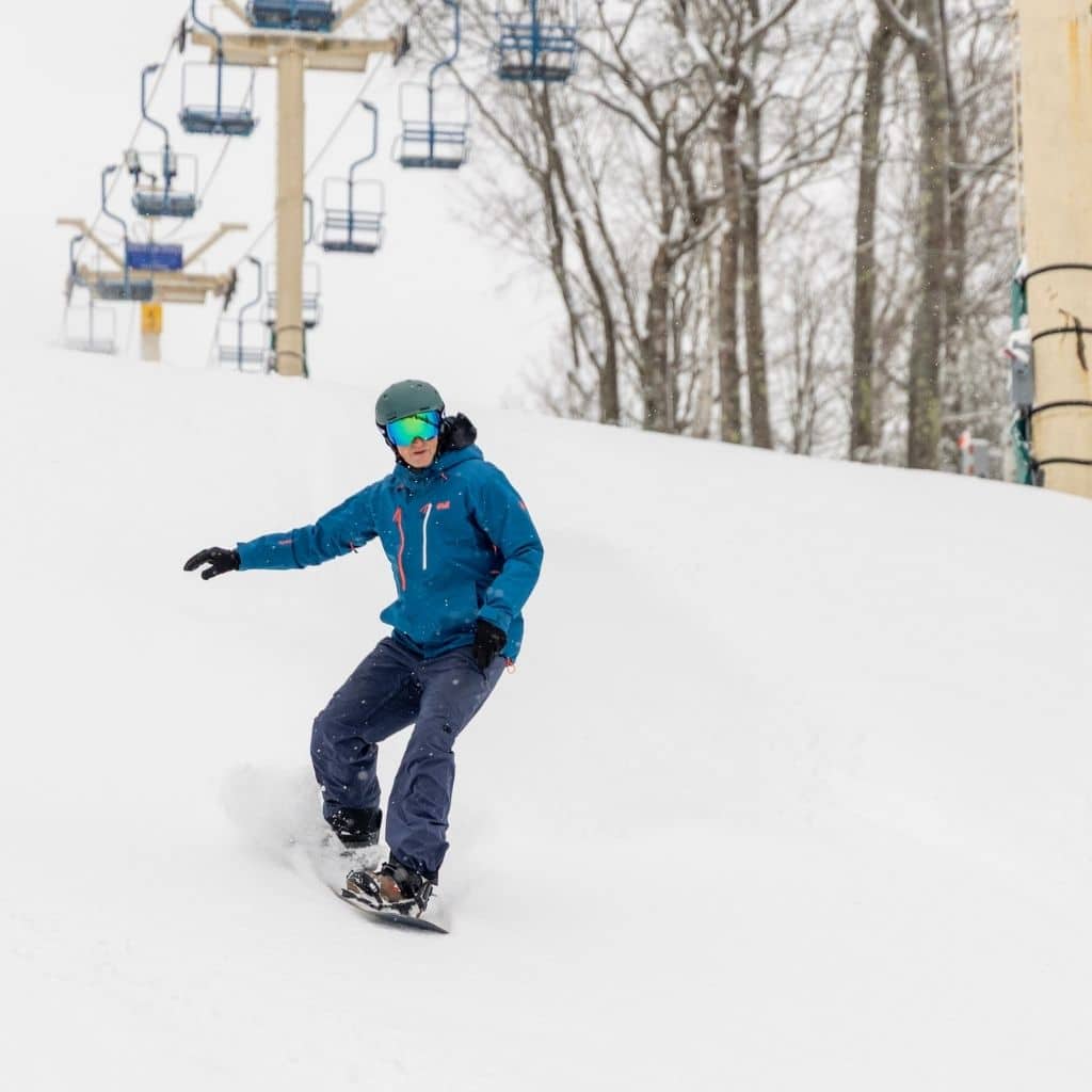 Snowboarder wearing a teal jacket and green helmet carves down a snow-covered slope with chairlifts in the background.