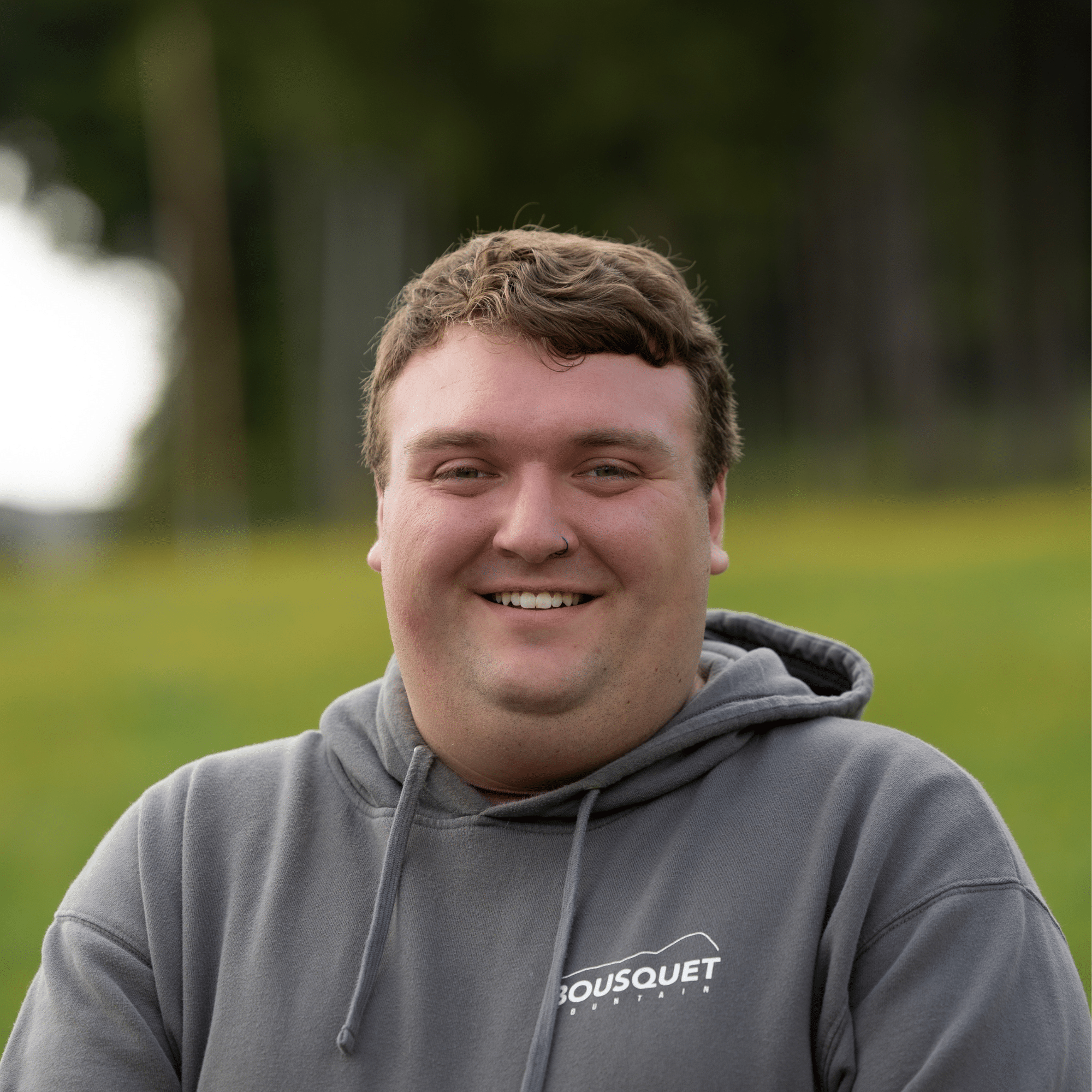 Smiling man in a gray hoodie outdoors, with a blurred green background; hoodie shows 'BOUSQUET' text.