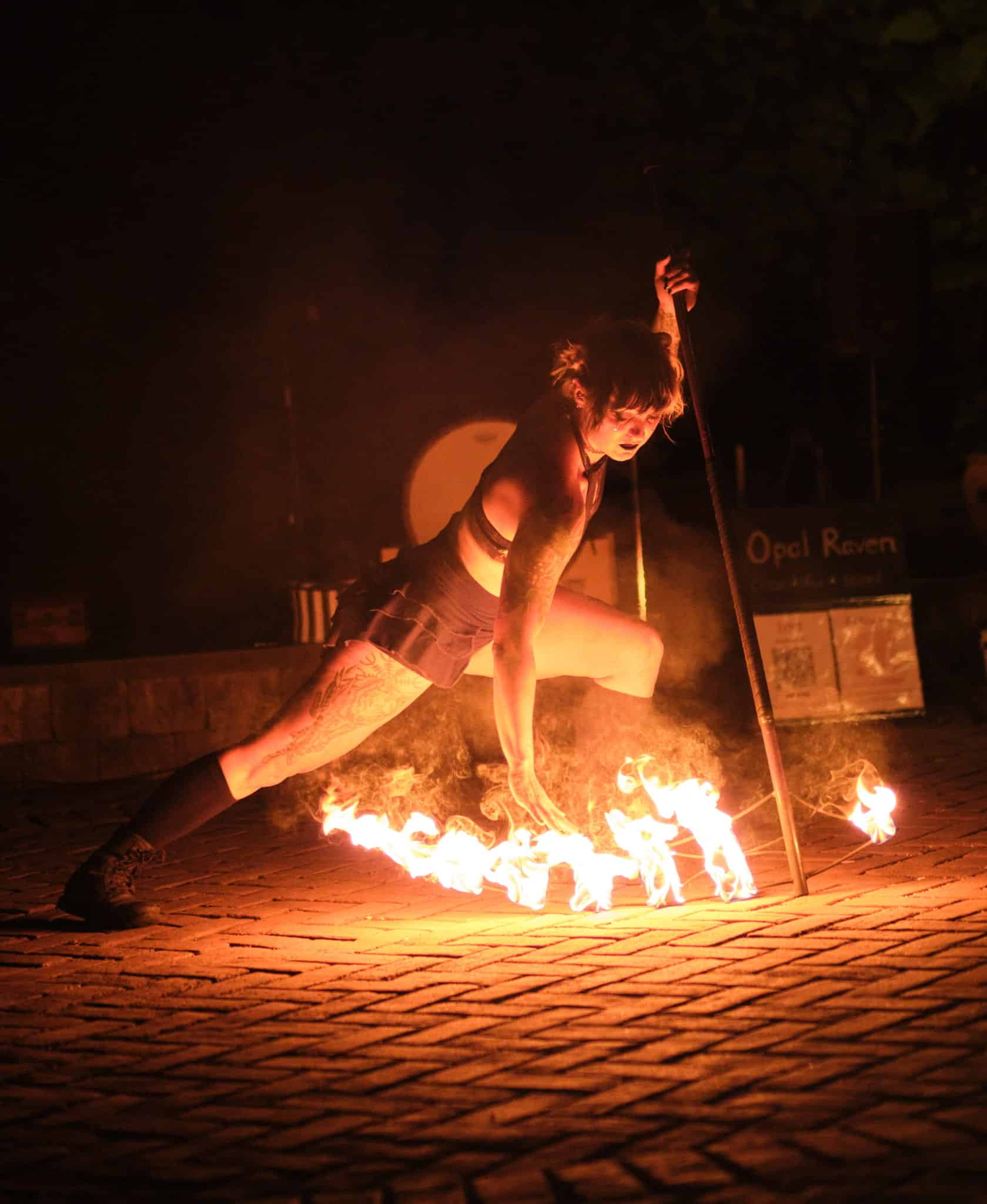 Nighttime fire performance: a tattooed performer crouches by flames, holding a burning torch in a dark outdoor setting