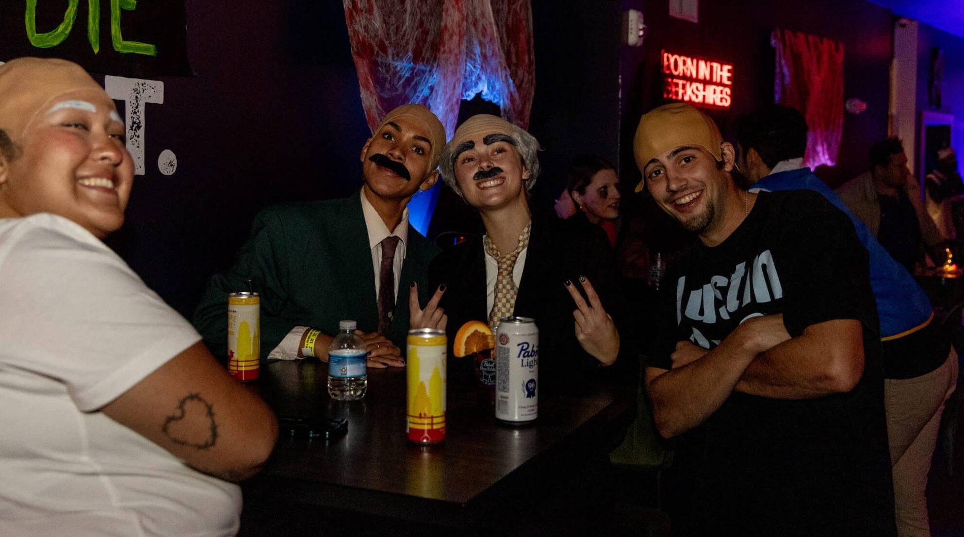Group of friends at a dimly lit party bar, posing for the camera with drinks, some in costumes and fake mustaches.